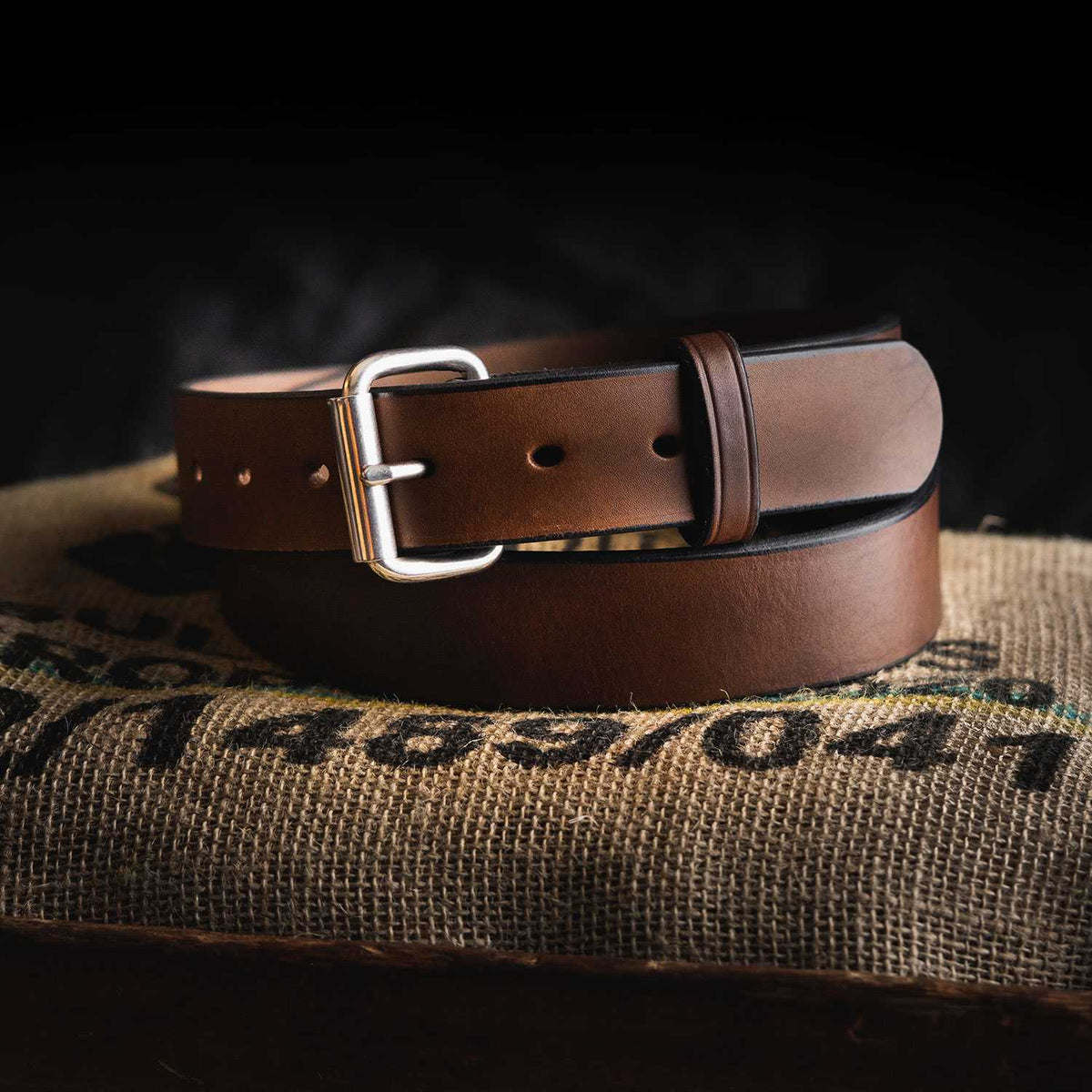 A Hanks Belts&#39; Gunner - 1.5&quot;, crafted from full-grain leather, is coiled neatly and placed on a burlap sack. The brown belt, USA made with a silver buckle and multiple holes for fastening, is set against a dark background.