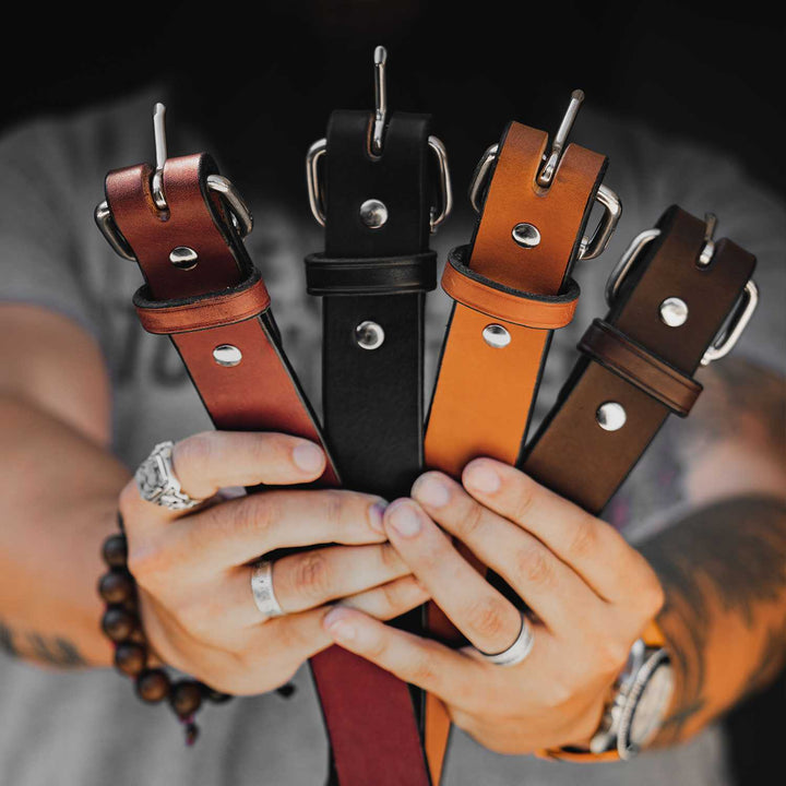 A person holds up four Hanks Belts "The Gunner - 1.5"" leather belts in different colors: red, black, light brown, and dark brown. Made from full-grain leather, these USA-made belts feature metal buckles and rivets. The person is wearing rings, a beaded bracelet, and a watch, with tattoos visible on their arms.