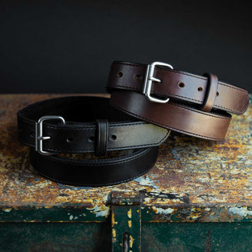 A close-up image of three Hanks Belts Kydex Reinforced CCW Belts - 1.5" placed on a rustic wooden surface. The belts come in different colors: black, brown, and tan. Each belt features a silver buckle and multiple adjustment holes, showcasing a polished and elegant design with vegetable-tanned leather.