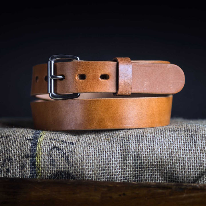 A close-up image of Hanks Belts' Old World Harness Leather Belt - 1.5" coiled on a burlap-covered surface. The belt, crafted from USA-tanned Hermann Oak leather, features a metal buckle with a pin and several adjustment holes. The dark background creates a striking contrast with the light-brown leather.