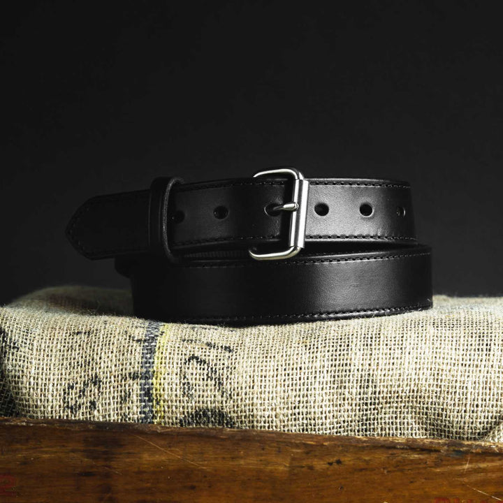 The Hanks Belts Kydex Reinforced CCW Belt - 1.5", crafted from black vegetable-tanned leather with a silver buckle, is coiled neatly on a burlap-covered surface against a dark background. This belt features multiple adjustment holes for sizing.