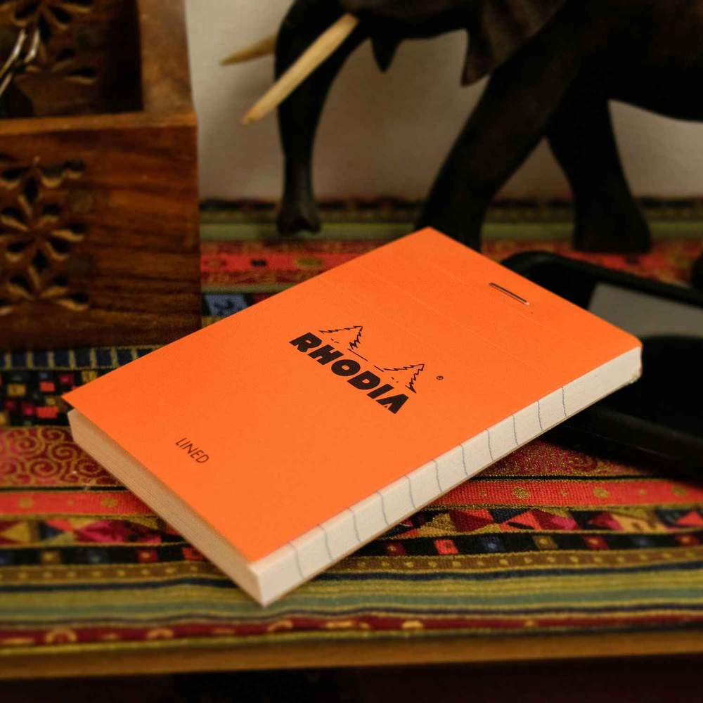 An orange Replacement Rhodia Notepad for Hanks Notesman lies on a colorful patterned surface. The notepad, known for its acid-free paper, is rectangular with a white spine. The cover displays the brand&#39;s logo with two mountain peaks. In the background, blurred objects include a wooden item and part of a black figure.