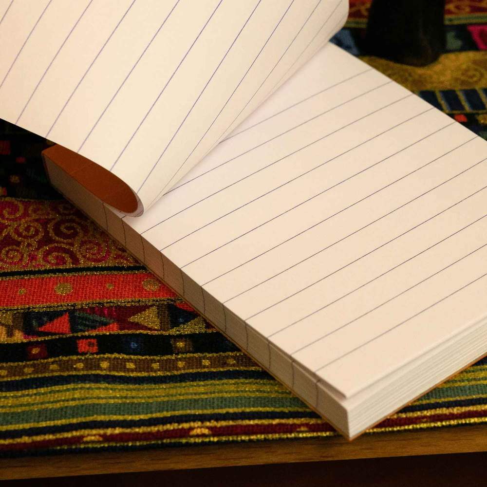 An open Replacement Rhodia Notepad for Hanks Notesman from Hanks Belts sits atop a colorful, patterned fabric. The notepad&#39;s acid-free pages are blank with evenly spaced blue lines. The vibrant fabric underneath features various geometric designs in red, green, yellow, and blue hues.
