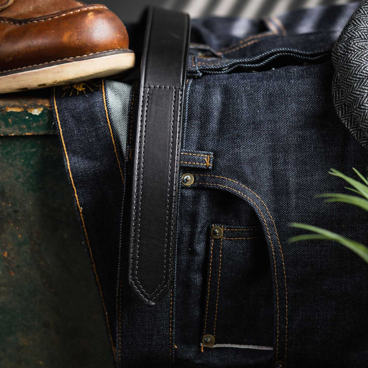 A pair of dark blue denim jeans with yellow stitching is laid on a green metal surface. A Scratchless Mechanics Belt - 1.5" by Hanks Belts, made of black leather, is looped through the belt loops of the jeans. A brown leather boot is partially visible at the top left, and a plant is in the bottom right corner.