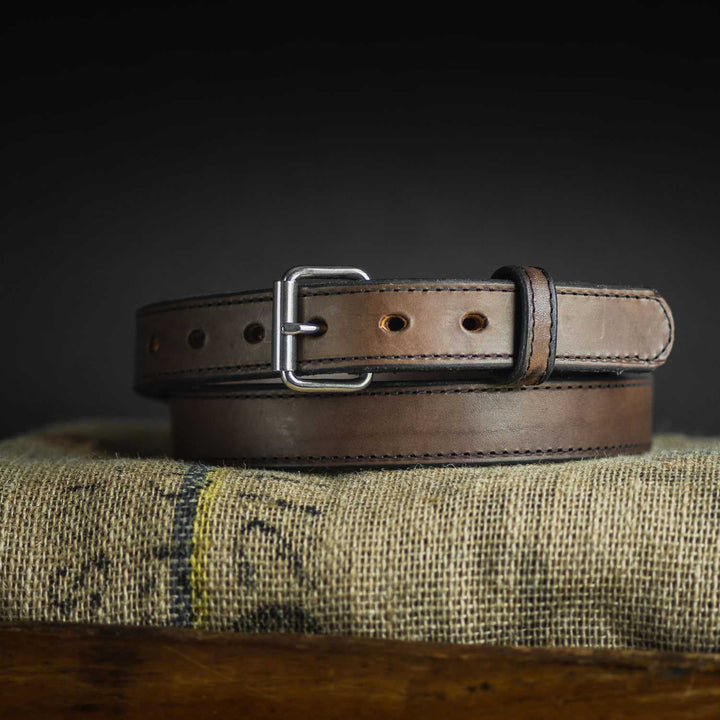 A Hanks Belts Steel Core Belt - 1.25", made from brown leather and featuring a silver buckle, is coiled neatly on a textured burlap fabric against a dark background. The extremely durable belt boasts multiple holes for adjustment and detailed stitching along the edges.