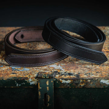 Two Hanks Belts' Scratchless Mechanics Belts - 1.5", one brown and one black, are coiled and displayed on a rusty, weathered metal surface. The close-up view highlights the belts' fine stitching and smooth, polished textures, emphasizing their quality craftsmanship in genuine full grain leather.