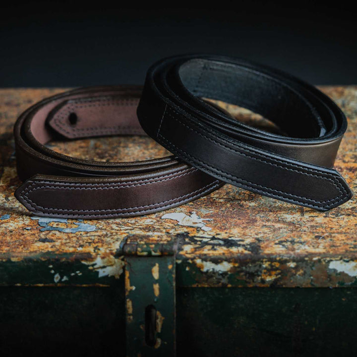 Two Hanks Belts' Scratchless Mechanics Belts - 1.5", one brown and one black, are coiled and displayed on a rusty, weathered metal surface. The close-up view highlights the belts' fine stitching and smooth, polished textures, emphasizing their quality craftsmanship in genuine full grain leather.