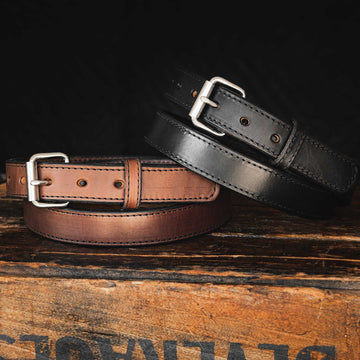 Two Hanks Belts' Xtreme Belts - 1.25" are displayed on a wooden surface. The brown belt on the left features a silver buckle, while the black belt on the right sports a durable heavy-duty roller buckle. Positioned against a dark background, these belts showcase exceptional craftsmanship, ideal for concealed carry use.