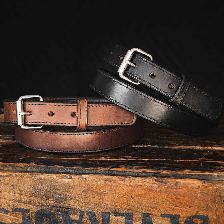 Two Hanks Belts' Xtreme Belts - 1.25" are displayed on a wooden surface. The brown belt on the left features a silver buckle, while the black belt on the right sports a durable heavy-duty roller buckle. Positioned against a dark background, these belts showcase exceptional craftsmanship, ideal for concealed carry use.