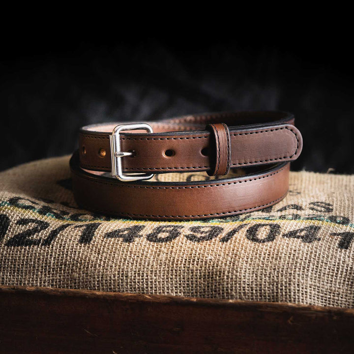 The Hanks Belts Xtreme Belt - 1.25" in brown leather, featuring a heavy-duty roller buckle, lies on textured burlap fabric with partial text and numbers visible. The dark background emphasizes the neatly coiled belt, highlighting the visible stitching along its edges.