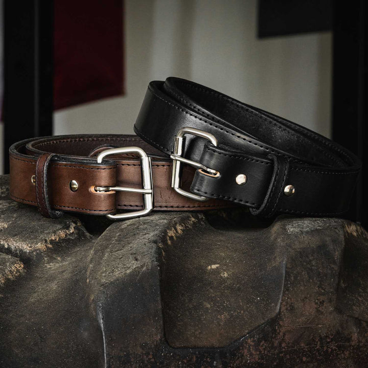 Close-up of two Hanks Belts Xtreme Belts - 1.75" draped over a textured stone surface. One belt is brown with a silver buckle, and the other is black with a silver buckle. Both heavy-duty leather belts feature double prong buckles and stitching details along the edges.
